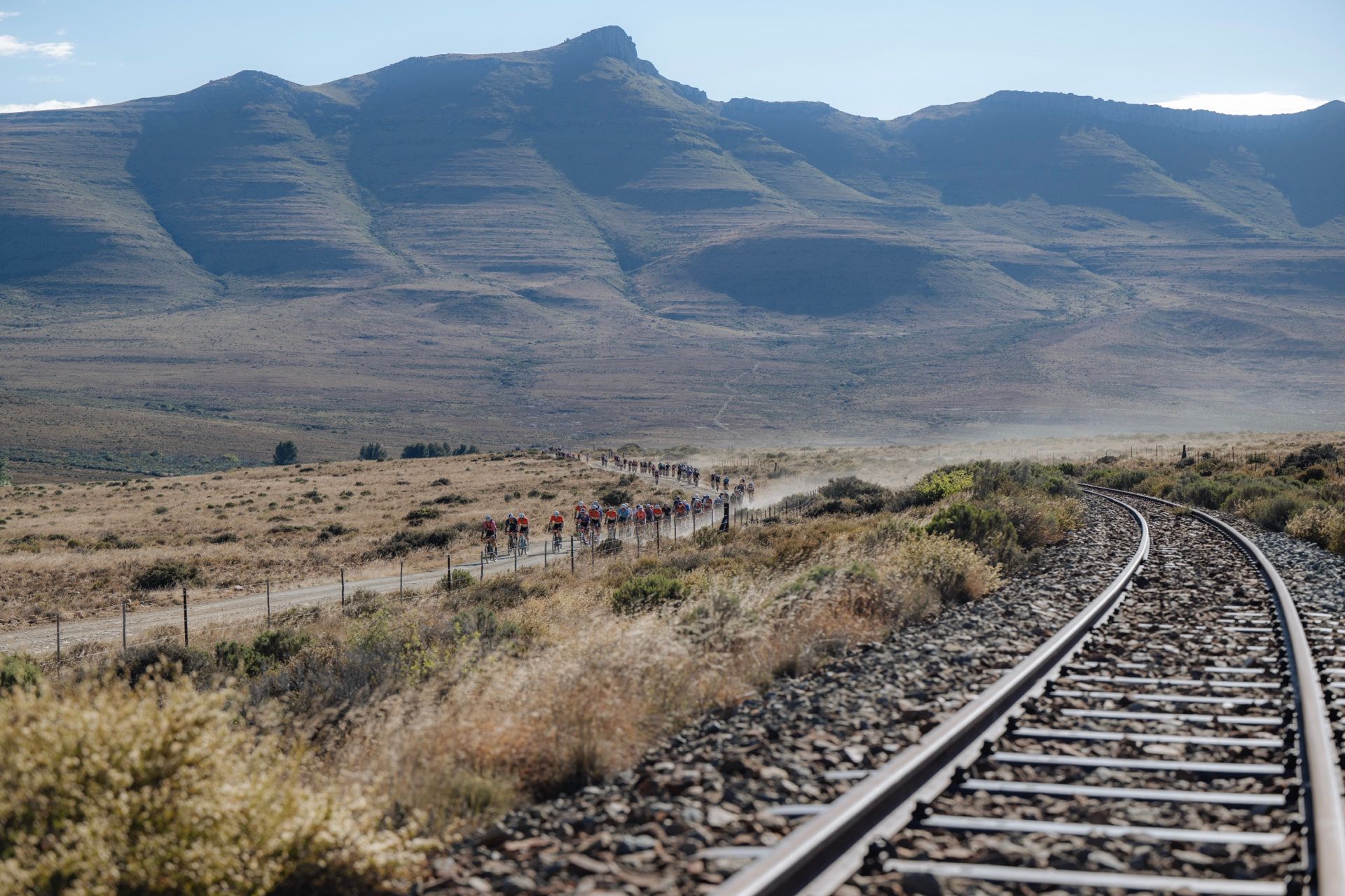 A long line of riders on a Karoo gravel road beside a railway line, under the mountains