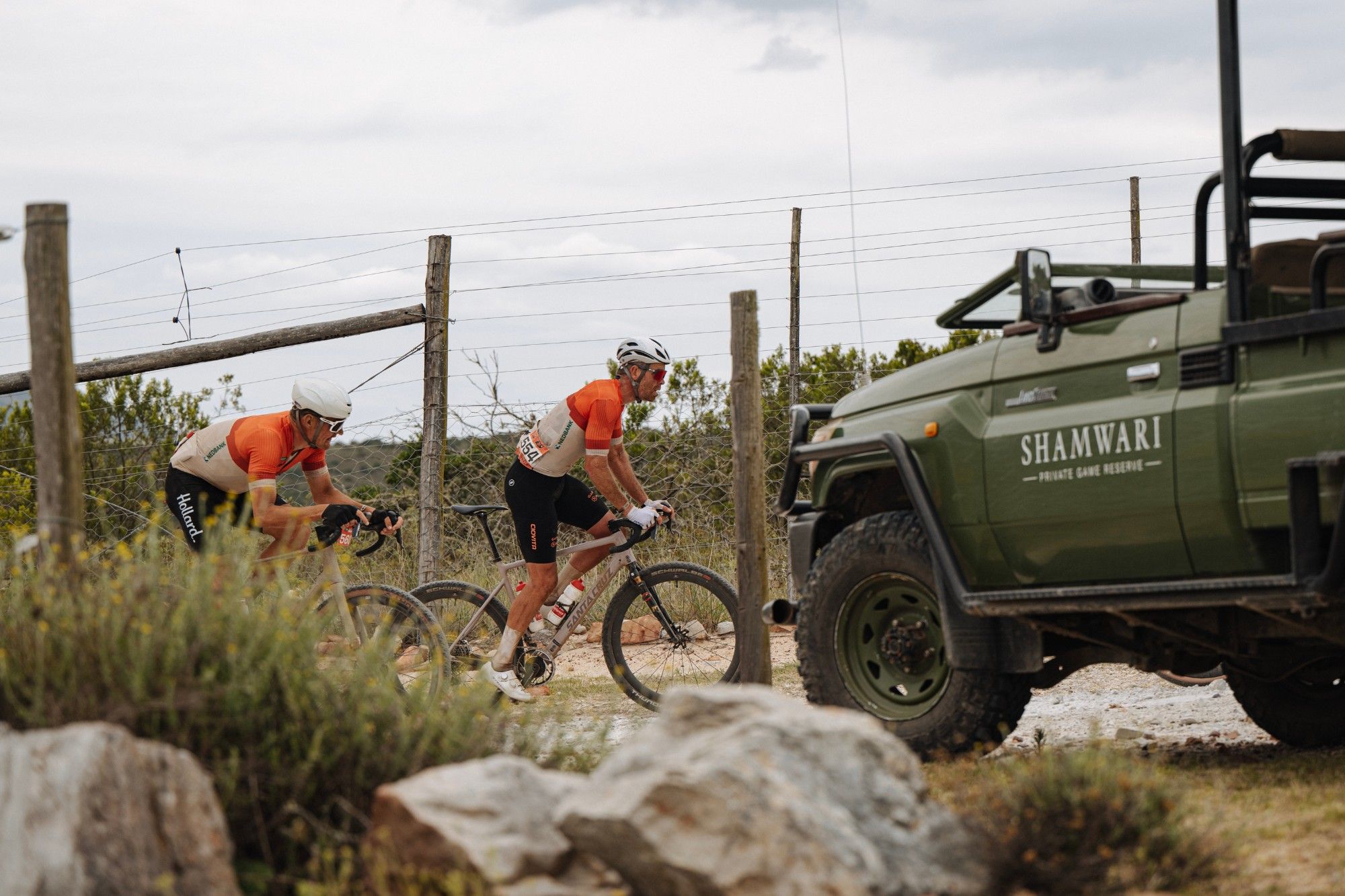 Two riders pass a Shamwari game-drive vehicle parked beside the gravel road