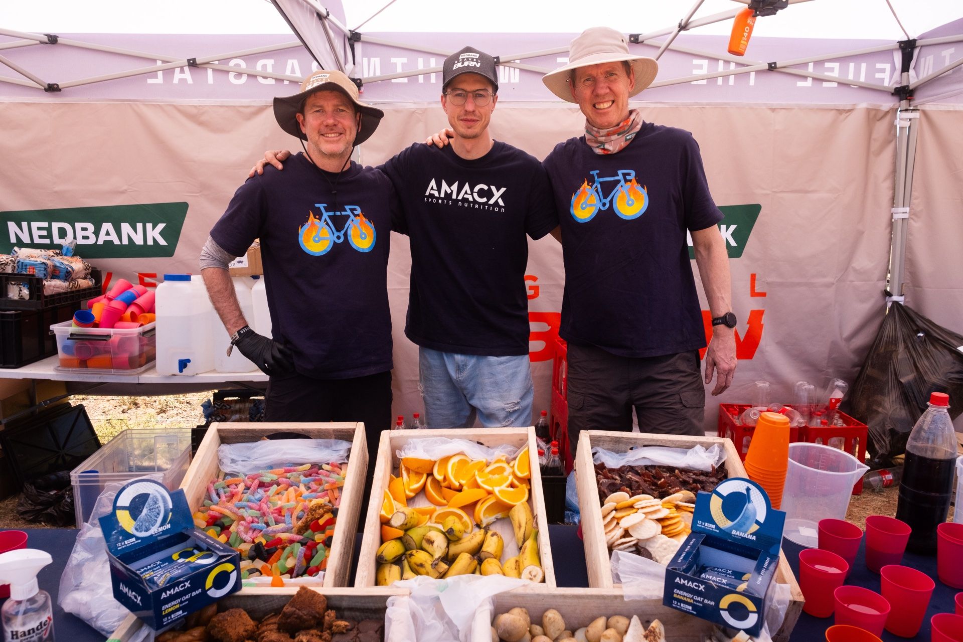 Three volunteers behind a Padstal table laid out with fruit, sweets and snacks